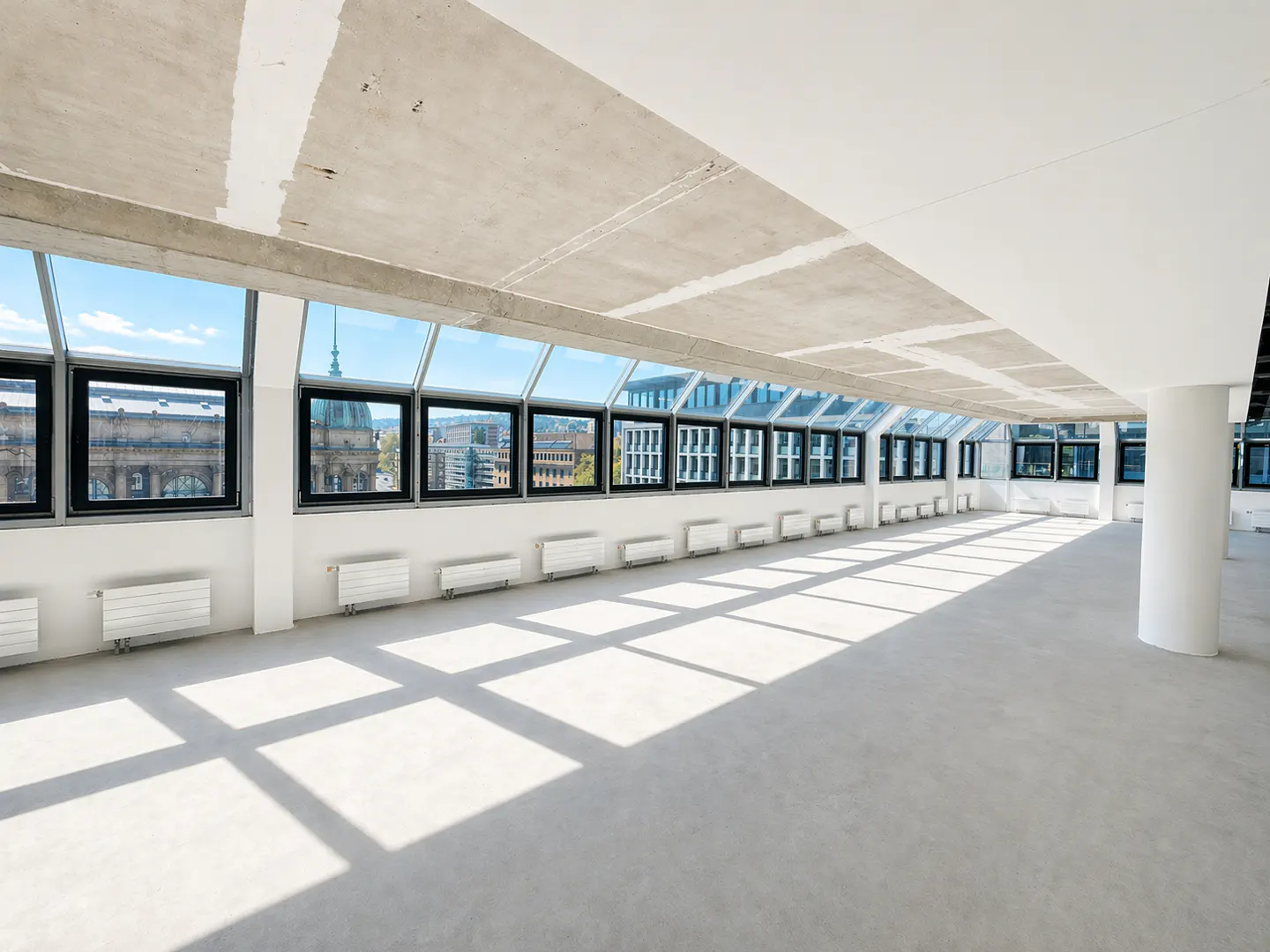 Bright, empty office space with concrete floors and ceiling. A wall of windows overlooks a city skyline.