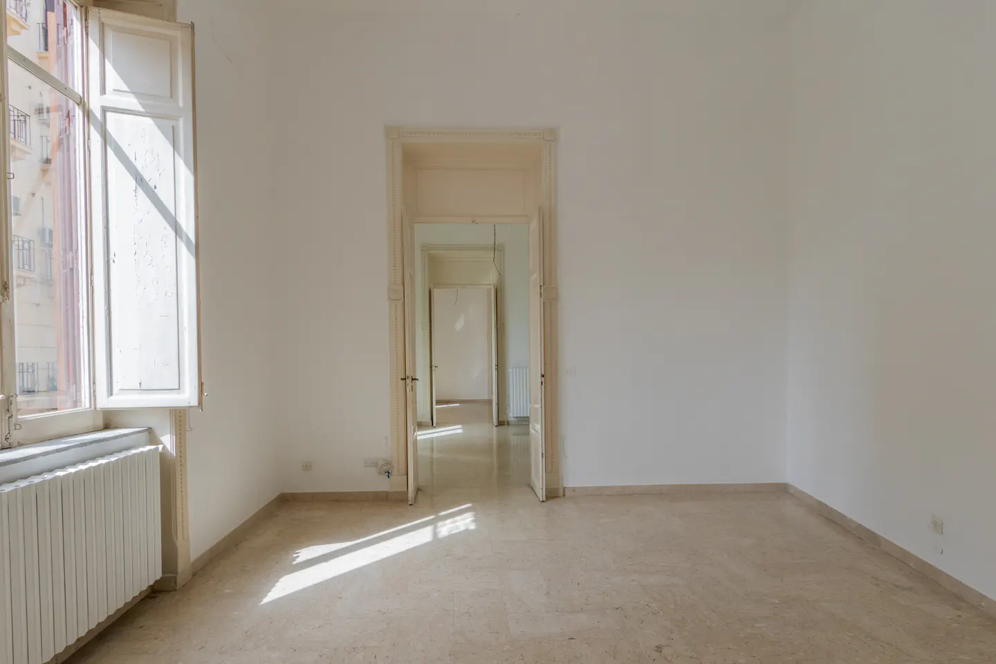 Bright, empty room with marble floors, white walls, and an open window with sunlight streaming in. A doorway leads to other rooms.