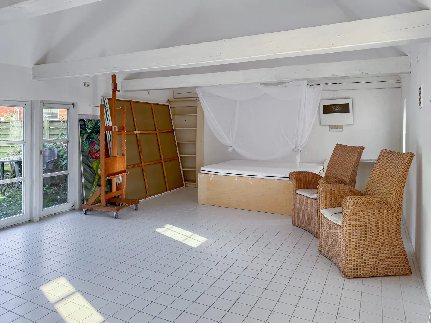 Bright, airy room with white tile floor, easel with painting, bed with canopy, and two wicker chairs.
