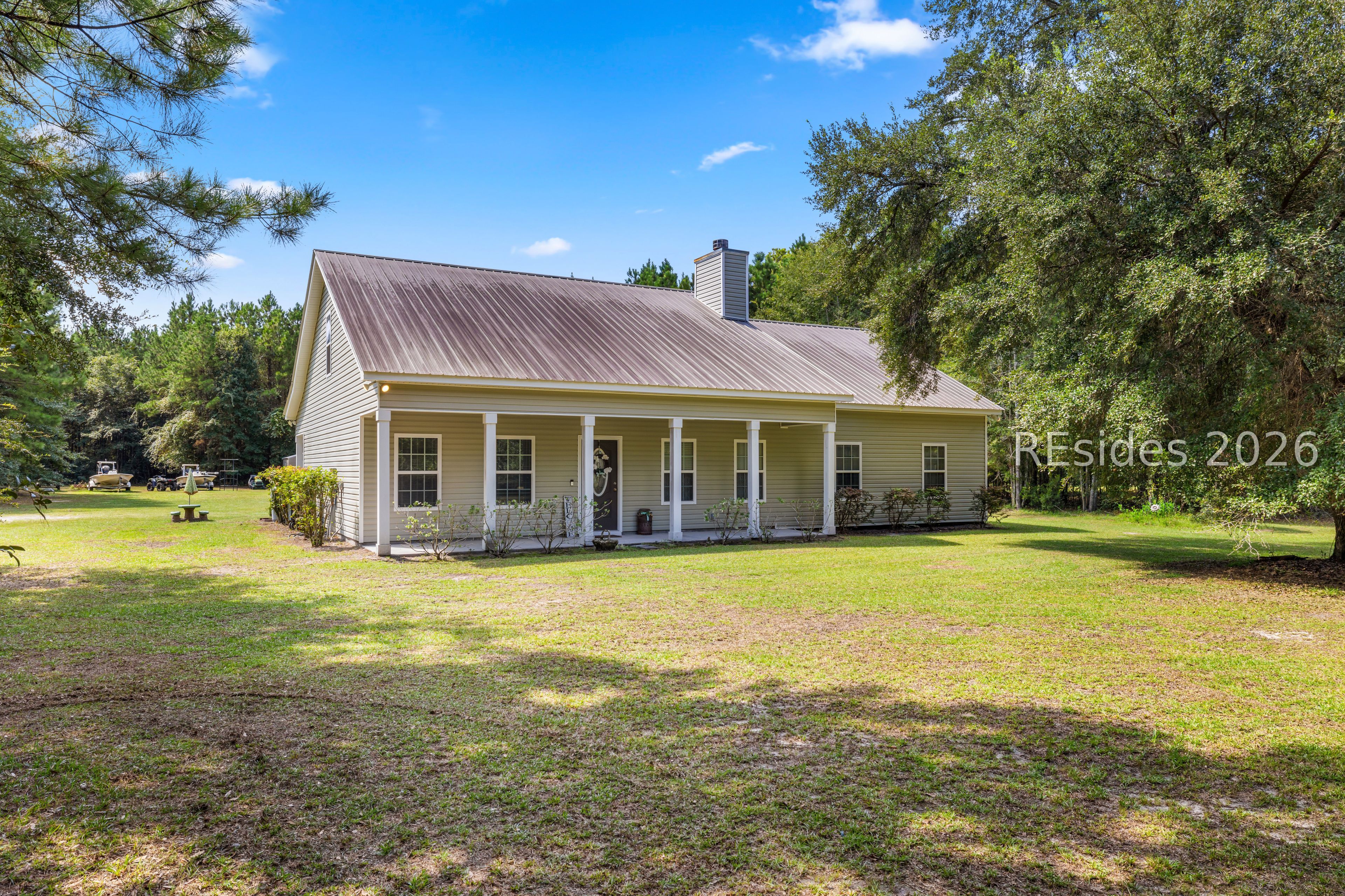 Single-story tan house with a metal roof and front porch, surrounded by a large grassy yard and mature trees. [AI-generated text]
