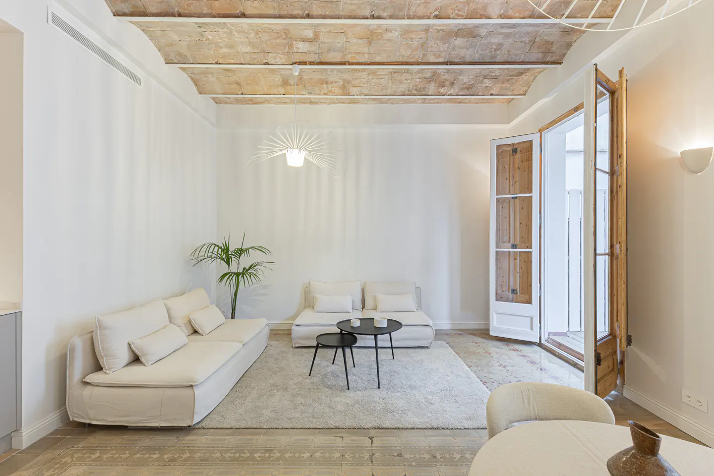 Bright living room with white sofas, black tables, and a palm. Open doors lead to a balcony. The ceiling has exposed brick.