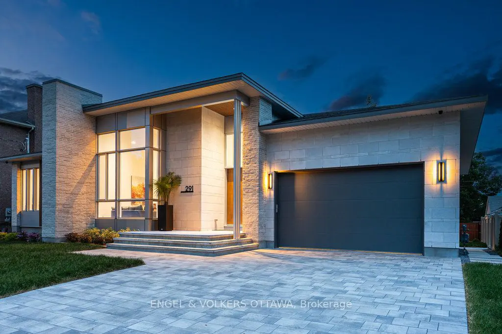 Modern, light-stone house with a gray garage door and brick driveway at dusk. Large windows reflect interior lighting.