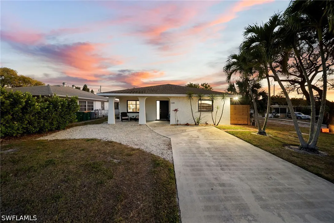 A white single-story house with a dark roof and a concrete driveway at sunset. Palm trees are on the right.