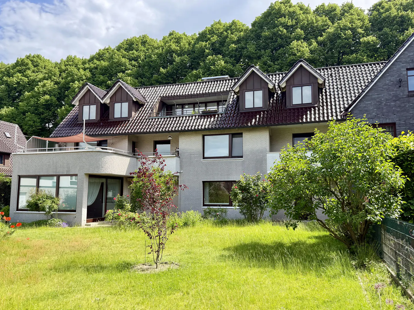 Two-story light brick house with a dark tiled roof, dormer windows, a balcony, and a lush green lawn against a forest backdrop.