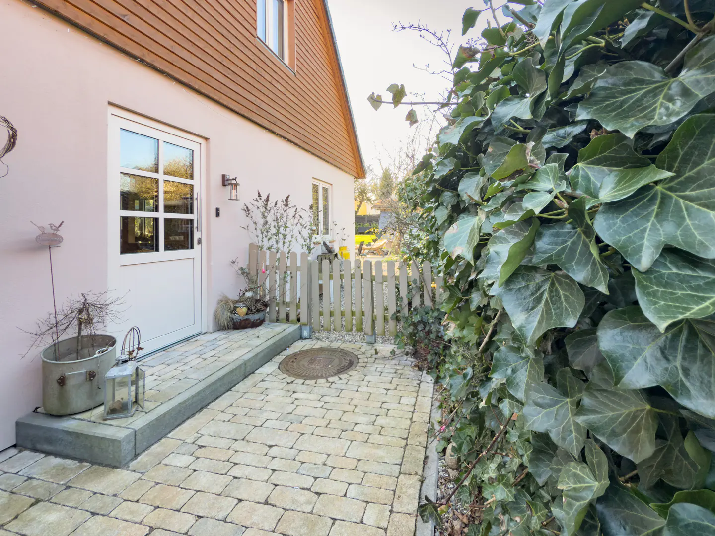 Exterior view of a house with a white door, brick walkway, and ivy-covered wall. A small picket fence and potted plants add charm.