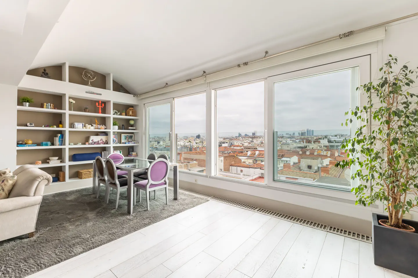 Bright room with white floors, a gray rug, and a glass table with purple chairs. Large windows overlook a city skyline. A bookshelf and potted plant add to the decor.