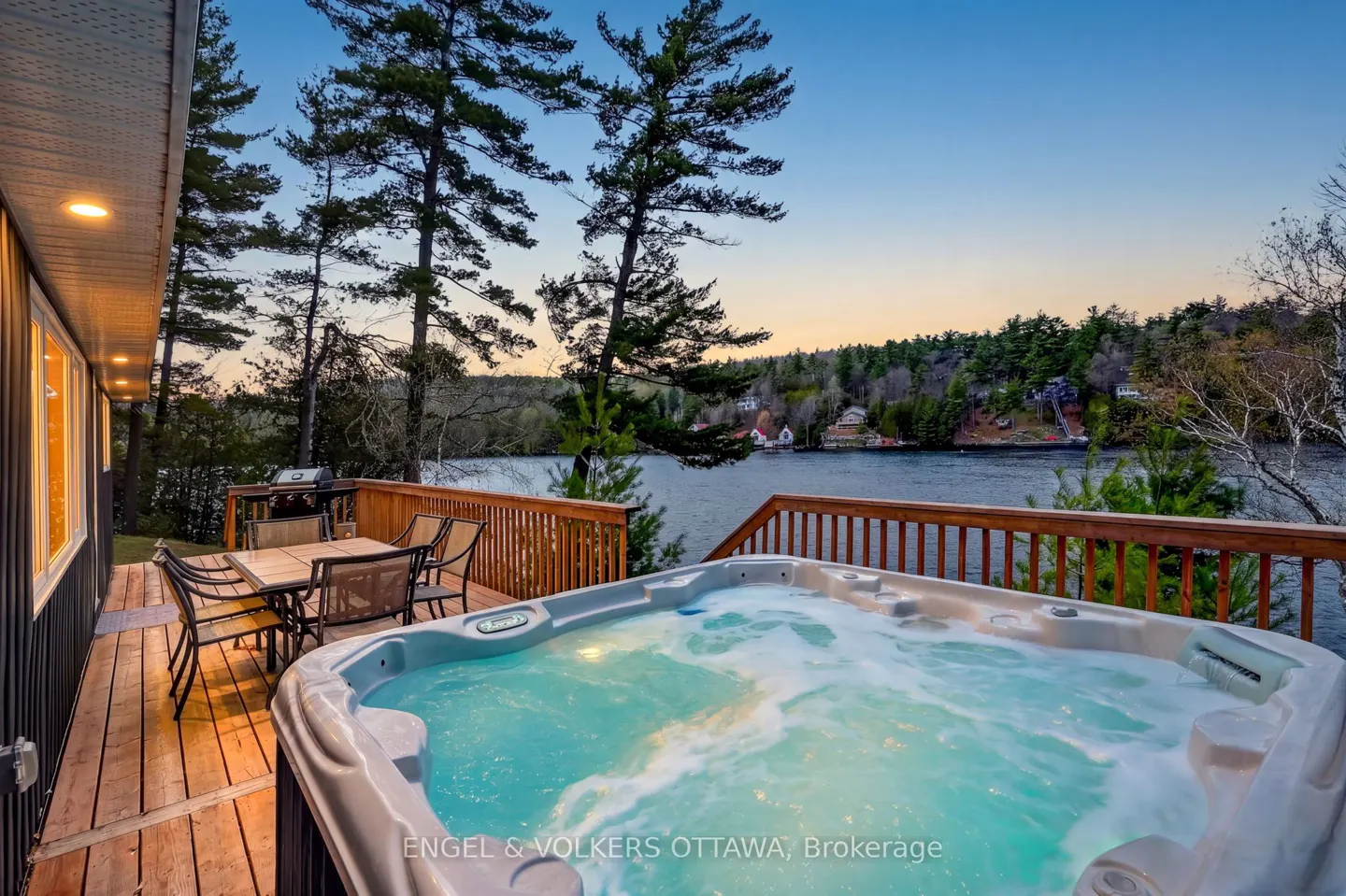 Outdoor deck with a hot tub overlooking a lake at dusk. Table, chairs, and grill are also on the deck. Tall trees surround the property.