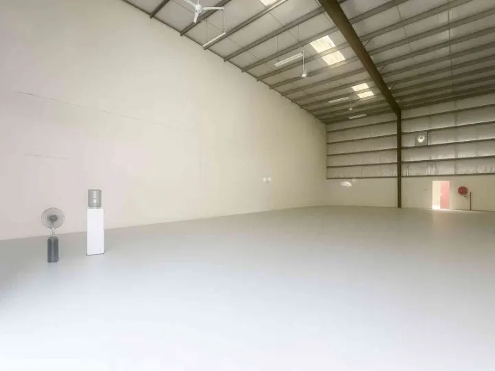 A bright, empty warehouse with white walls, a metal roof, and a gray floor. A fan and water cooler sit in the corner.