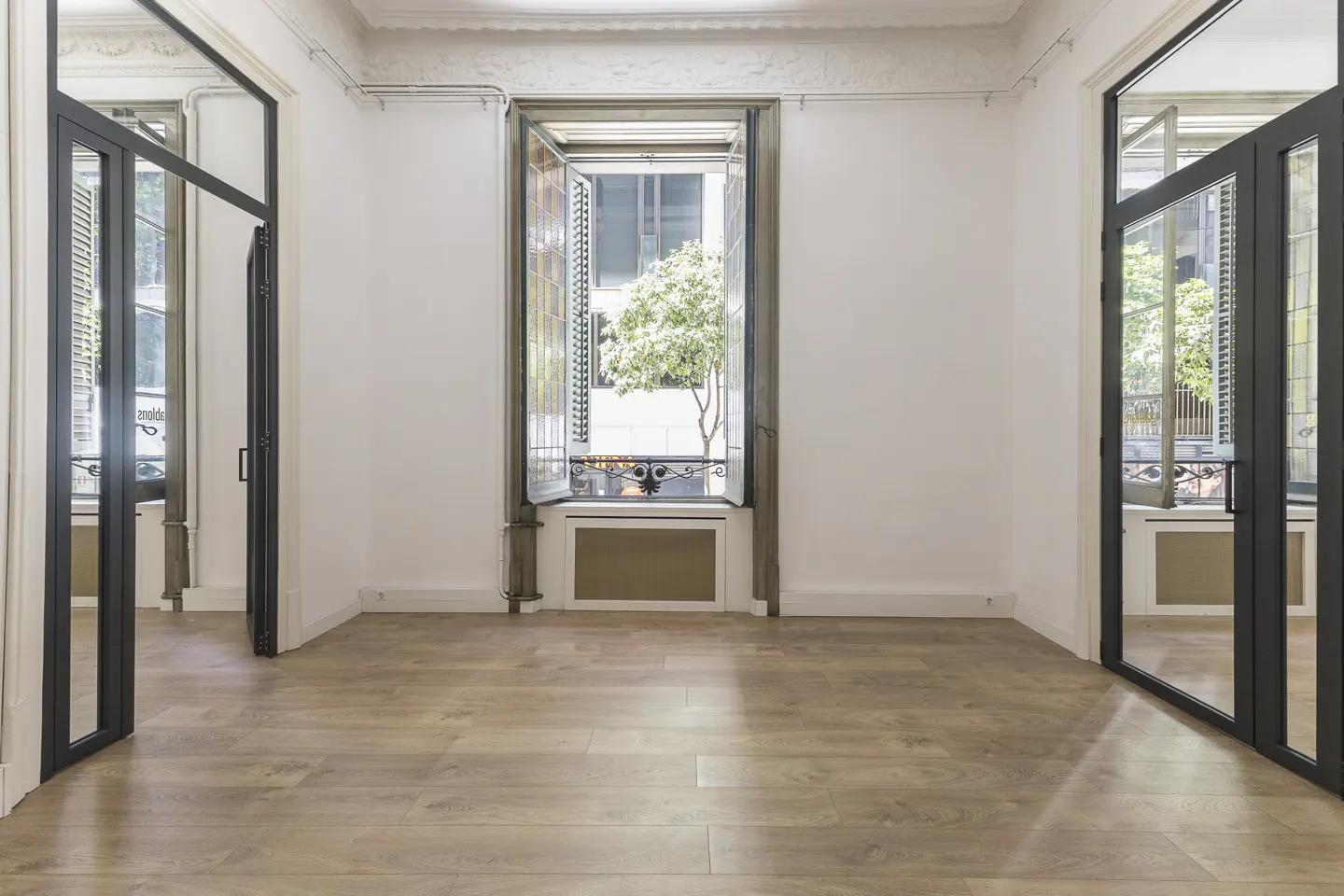 Empty room with light wood floors, white walls, and black framed glass doors. An open window shows a tree outside.