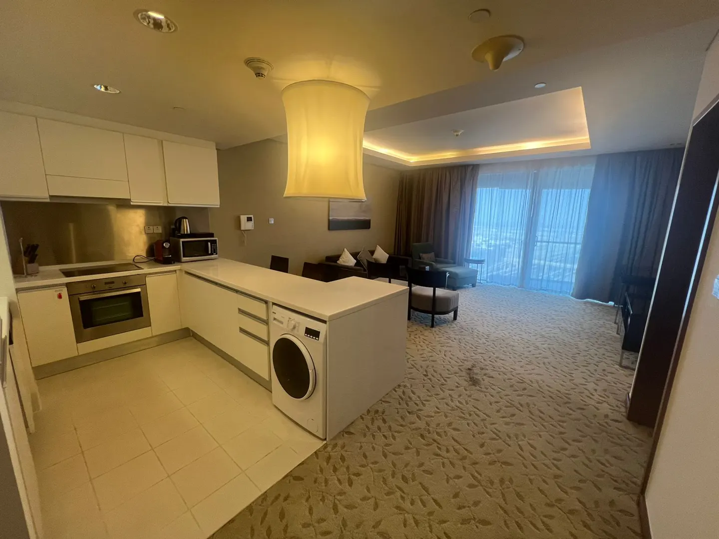 A wide shot of a modern apartment interior with white kitchen, living area, and patterned carpet. Curtains frame a bright window view.