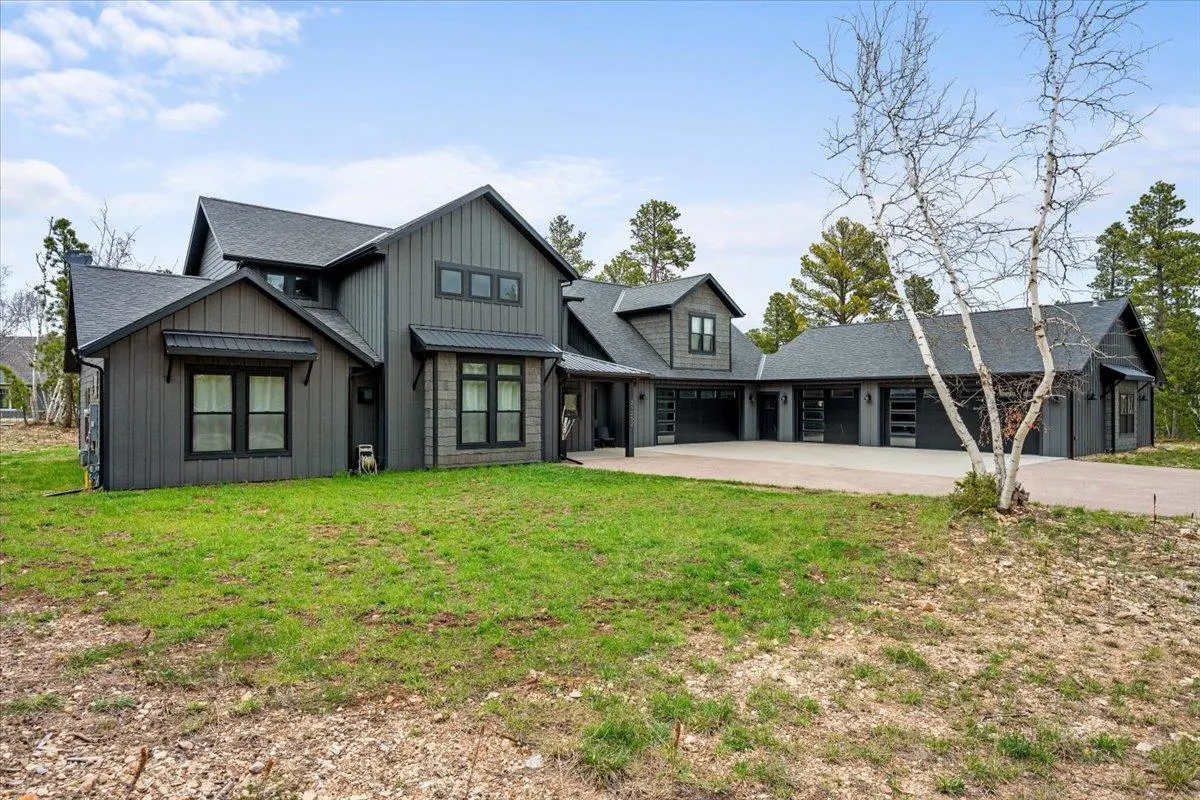 Modern dark gray farmhouse with a large concrete driveway, multi-car garage, and surrounding forest under a blue sky.