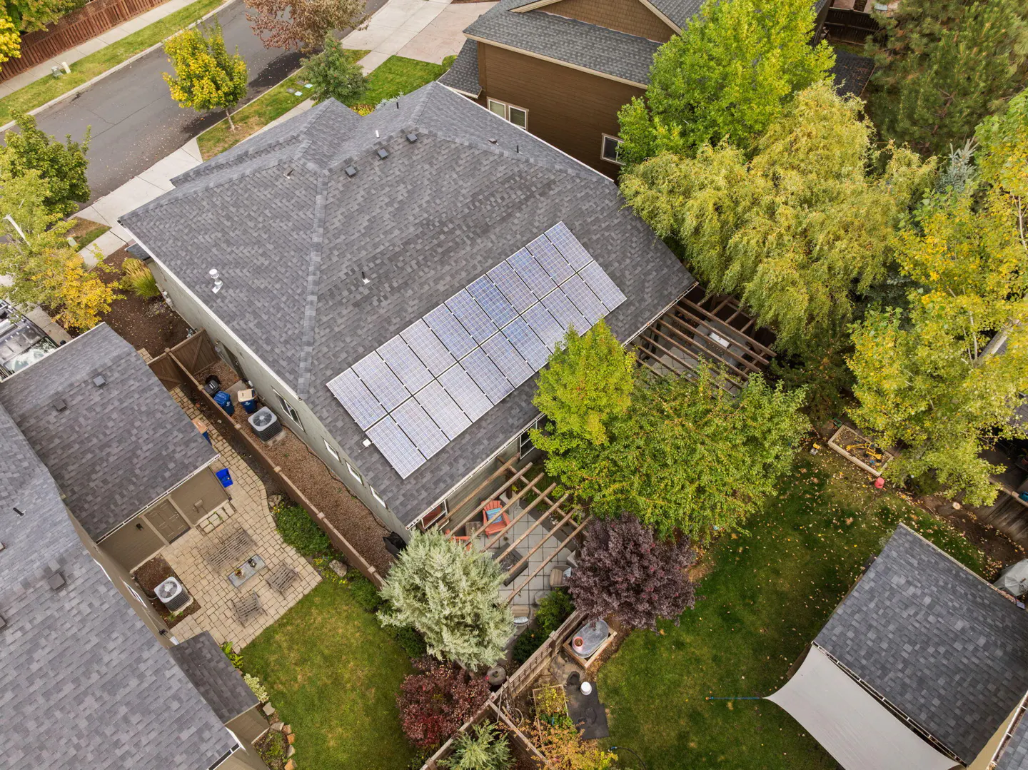 Aerial view of a gray house with solar panels on the roof, a wooden pergola, and a green lawn.