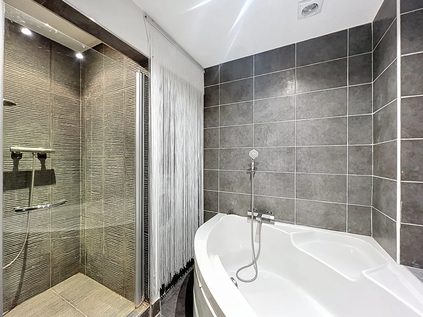 Bathroom with gray tiled walls, a white bathtub with a shower head, and a glass-enclosed shower with a silver shower fixture.