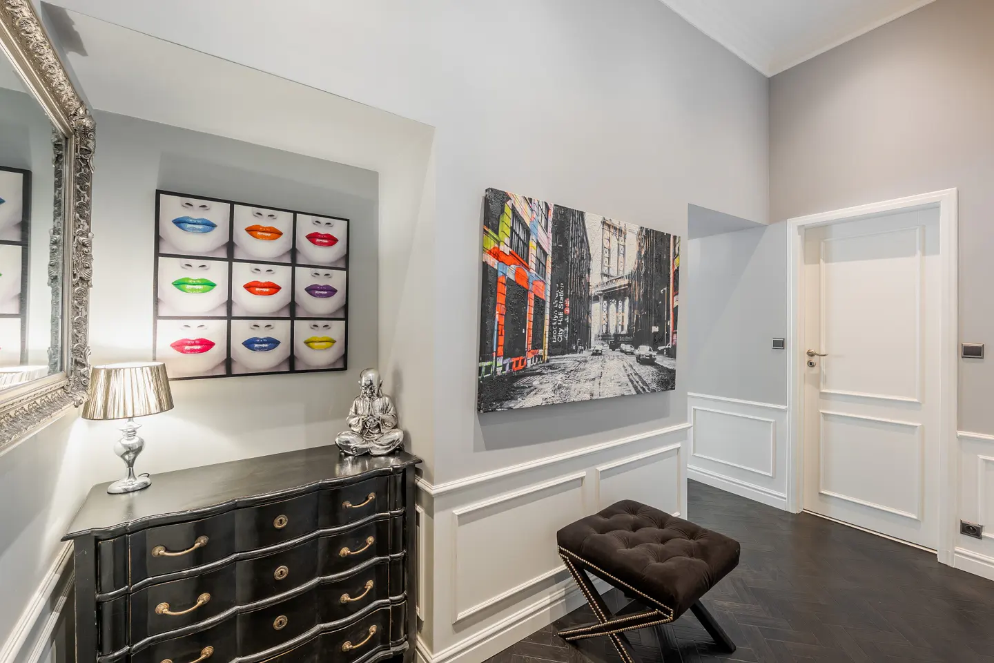 Hallway with gray walls, dark wood floors, and white trim. A black dresser with gold pulls sits under a colorful lip art print and a Buddha statue. A city painting hangs above a brown tufted stool.