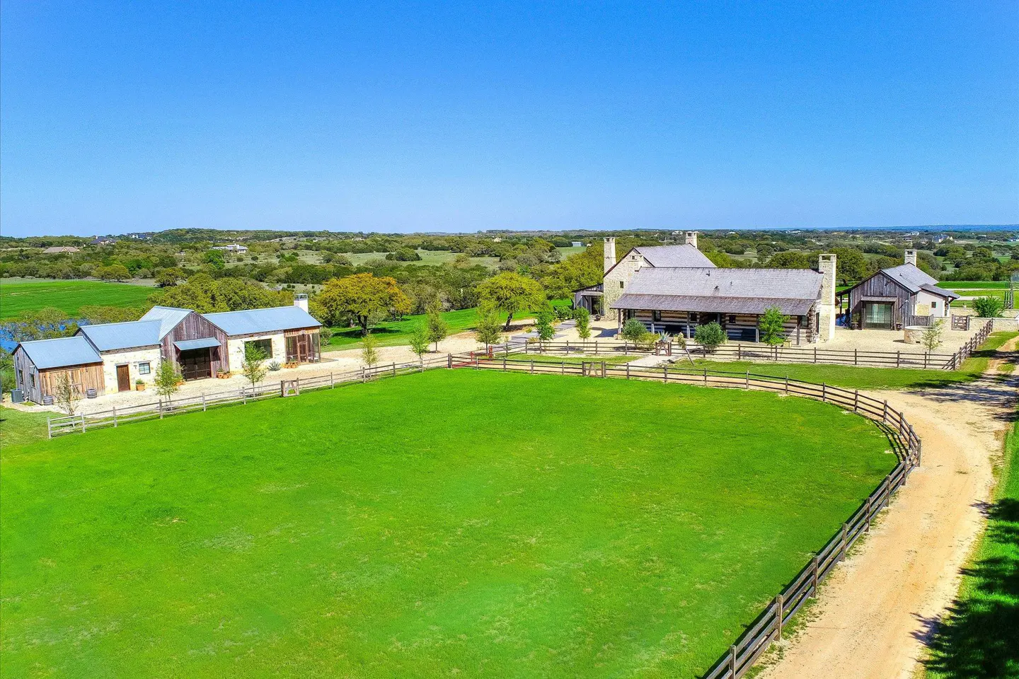 Aerial view of a ranch with a large green field, wooden fence, and several stone and wood buildings under a clear blue sky.