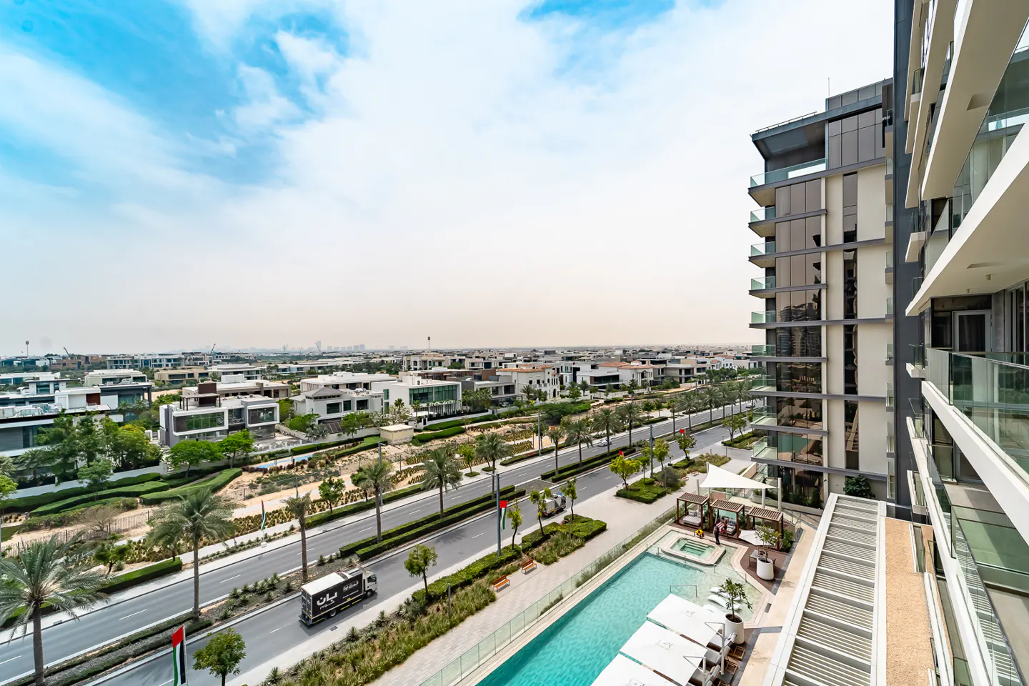 View from a high-rise balcony overlooking a pool, palm tree-lined street, and modern homes under a blue sky.