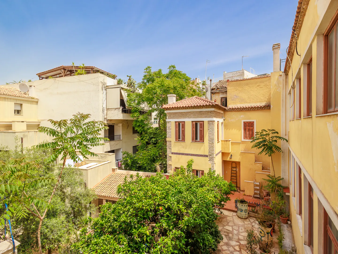 View of yellow buildings with red-tiled roofs, green trees, and a stone courtyard under a blue sky.