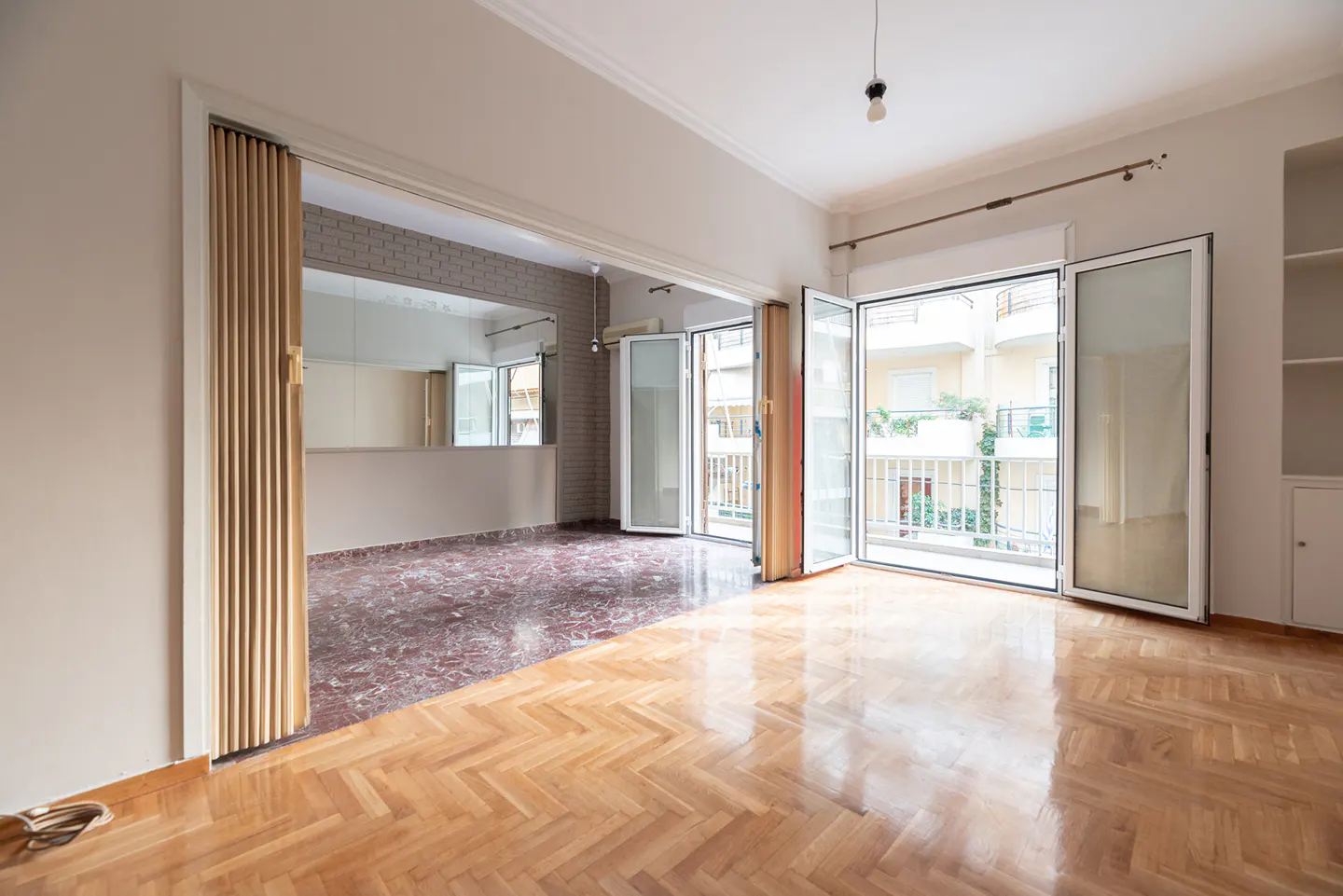 Bright, empty room with herringbone wood floors and open doors to a balcony. A second room with marble floors is visible.