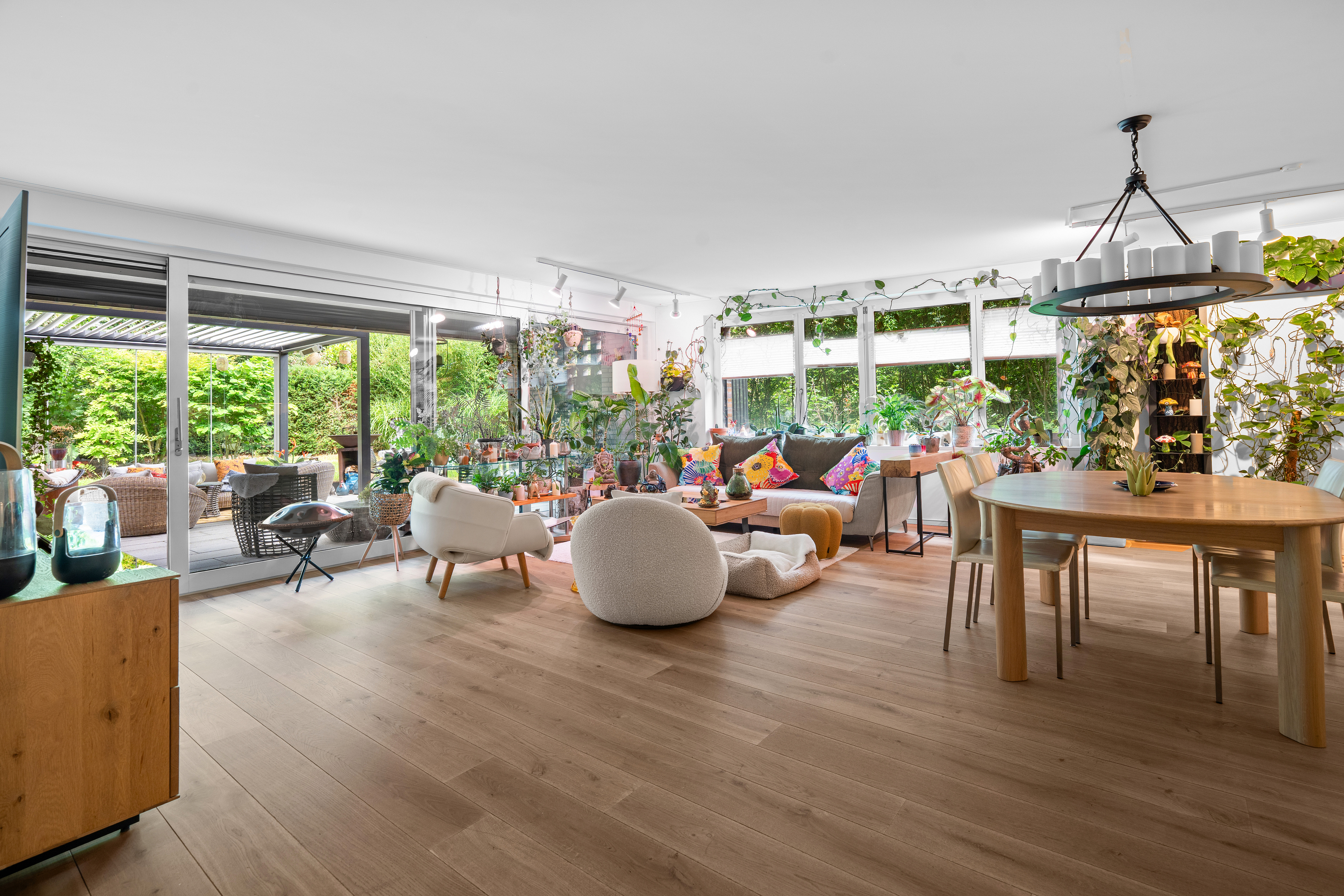 Bright, open-plan living room with light wood floors, modern furniture, many indoor plants, and a large sliding glass door.