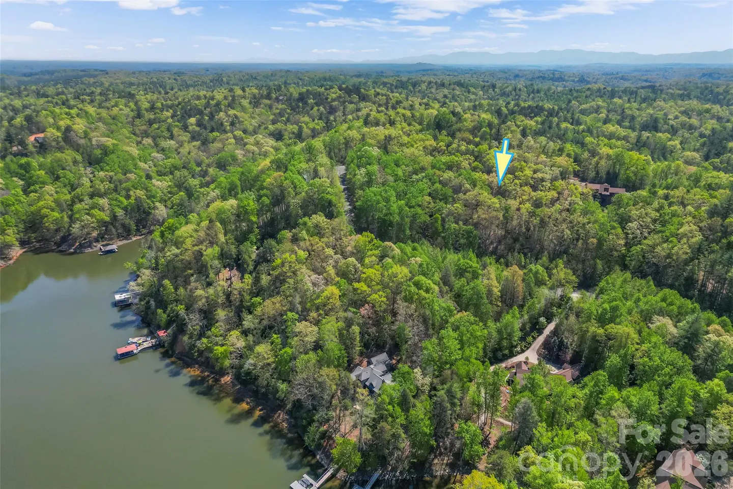 Aerial view of wooded real estate lot for sale, near a lake with docks. A blue arrow points to the property.