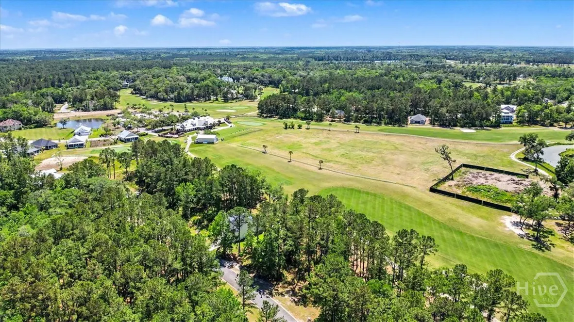 Aerial view of a lush green golf course surrounded by dense trees and scattered houses under a blue sky.
