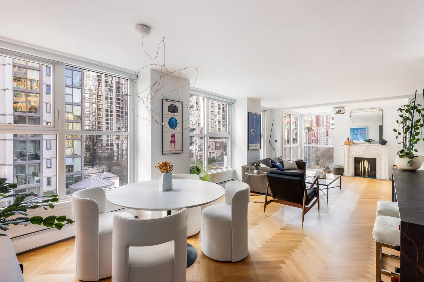 Bright, modern apartment interior with herringbone wood floors. A white round table and chairs sit near large windows with city views. A fireplace is visible in the living area.