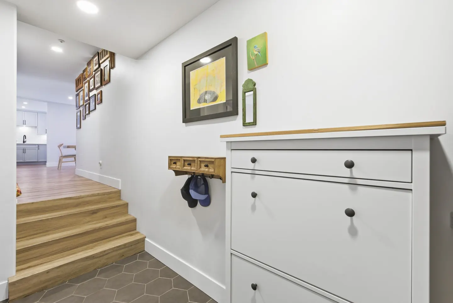 Entryway with white walls, brown hexagonal tile floor, and wooden stairs. A white cabinet and wall art decorate the space.