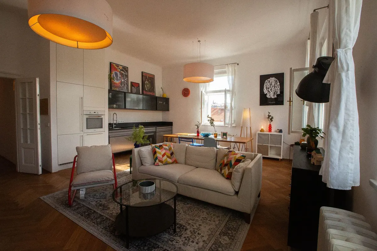 Bright, open-concept apartment with a cream sofa, glass coffee table, and a red-framed chair on a patterned rug. A kitchen and dining area are in the background.