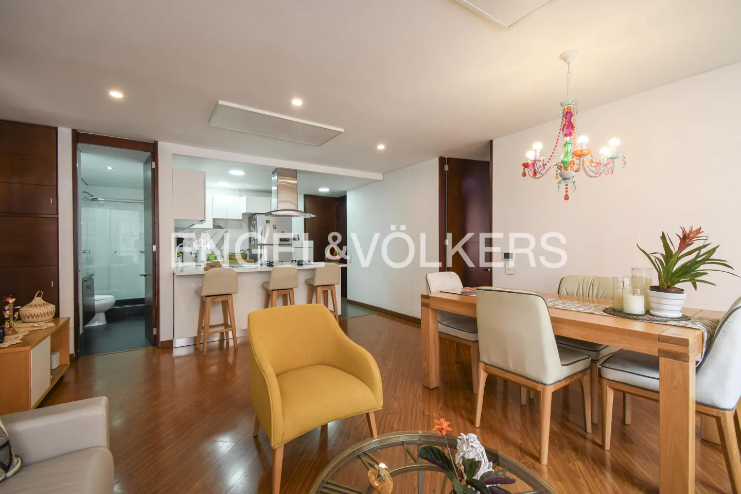 Open-concept living space with wood floors, a yellow chair, a dining table with four chairs, and a kitchen with bar stools. A colorful chandelier hangs above the dining table.