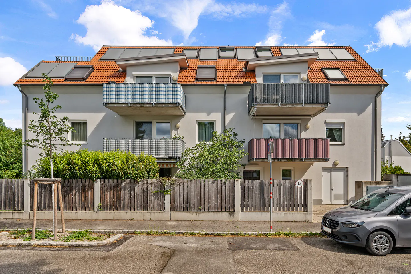 Three-story apartment building with balconies, orange roof, solar panels, and a gray car parked in front.
