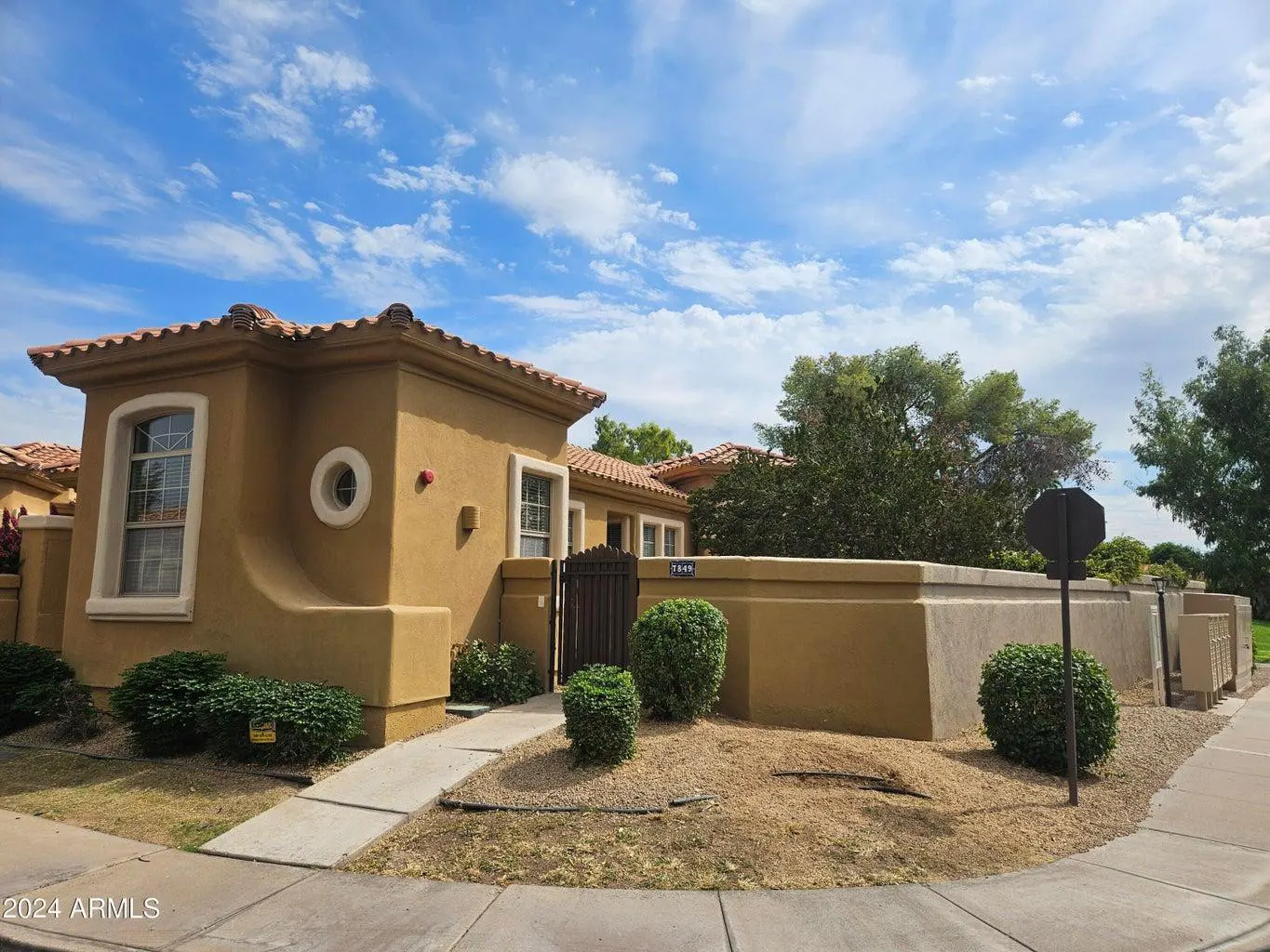 Tan stucco home with a red tile roof, small yard, and blue sky with white clouds.
