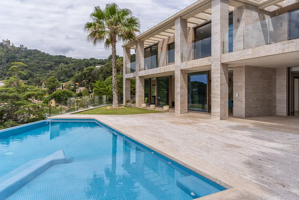 Luxury home with a blue tiled pool, stone patio, and a palm tree. A green hillside is in the background.