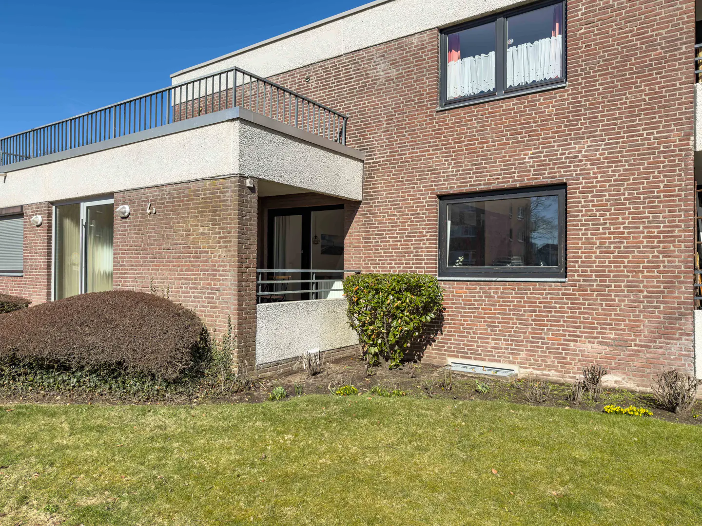 Exterior view of a two-story brick apartment building with a balcony, green lawn, and blue sky.