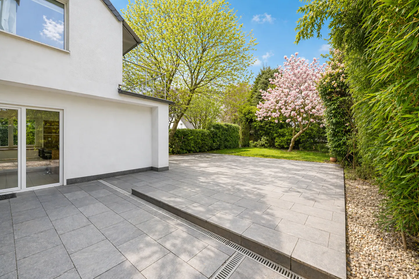 Exterior view of a modern white house with a gray stone patio, green trees, and a pink magnolia tree.