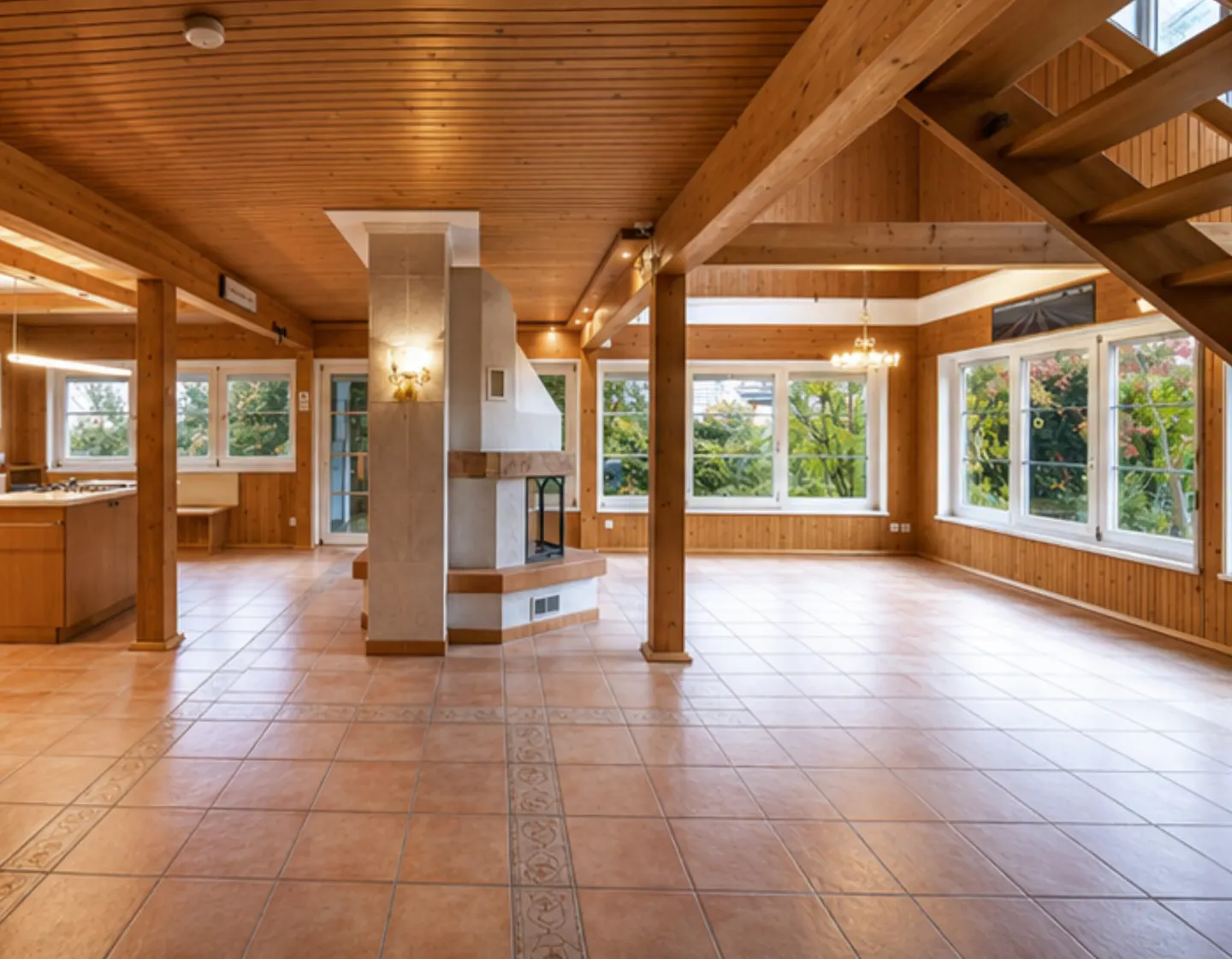 Spacious living room with wood-paneled walls, tile floors, and a central fireplace. Large windows offer views of greenery.