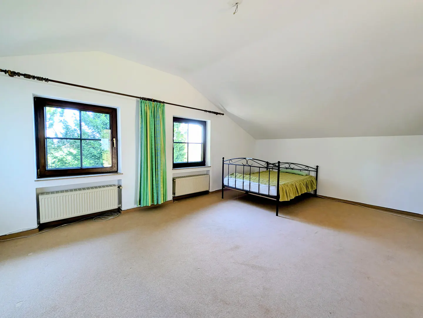 Attic bedroom with white walls, carpet, and a metal bed frame with a green bedspread. Two windows with green curtains let in natural light.