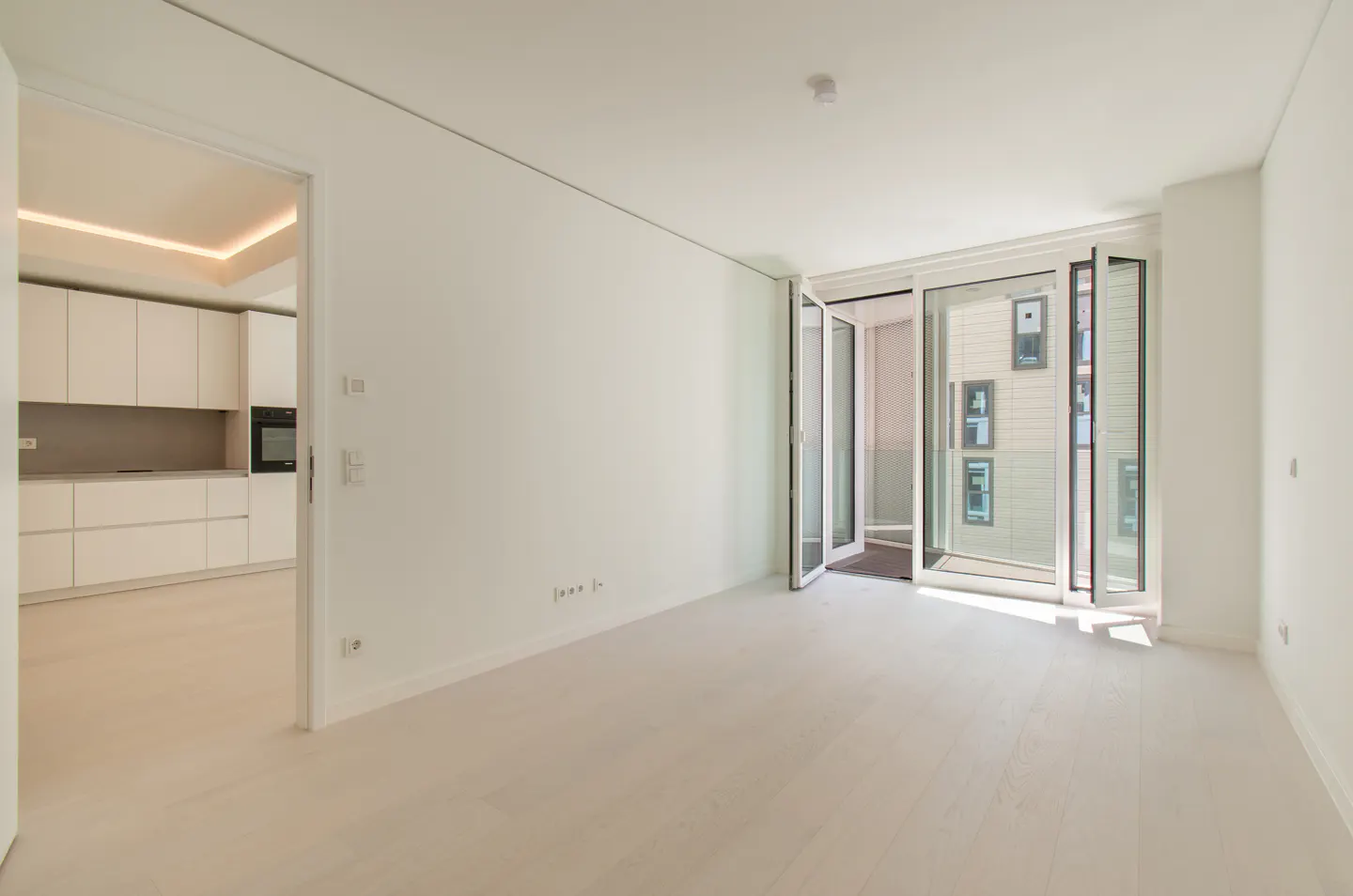 Bright, empty room with white walls and light wood floors. Open glass doors lead to a balcony. A kitchen is visible through a doorway.