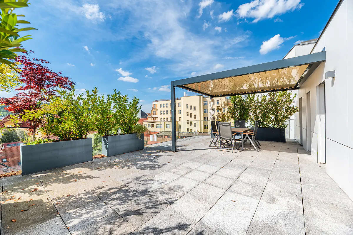 A rooftop patio with gray stone tiles, planters with greenery, and a pergola over a table and chairs. City buildings are visible in the background.