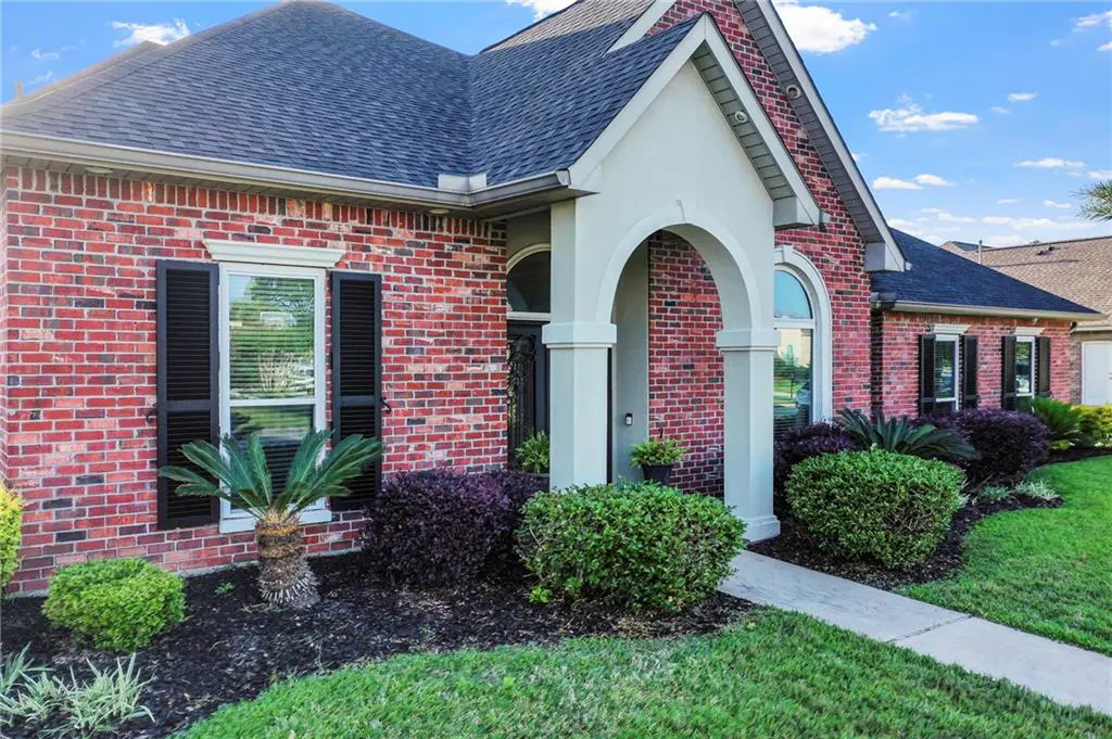 Red brick house with black shutters, a dark roof, and a white arched entryway. Green bushes and grass surround the house.