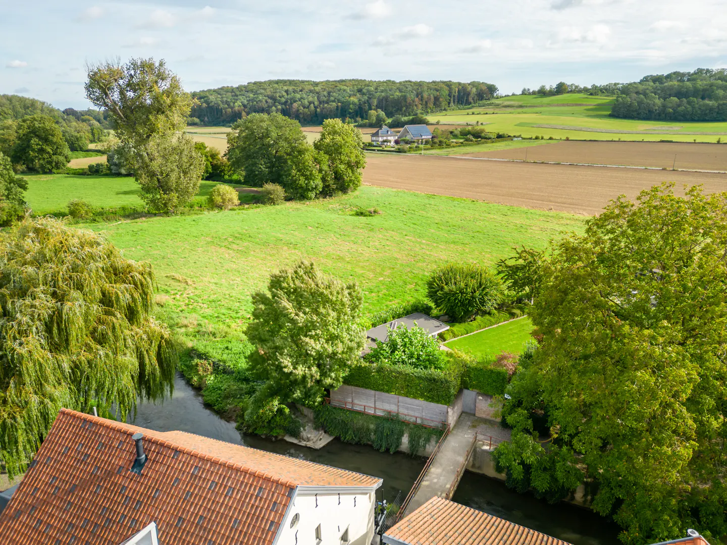 Aerial view of a rural landscape with green fields, trees, a river, and a house with a red tile roof.