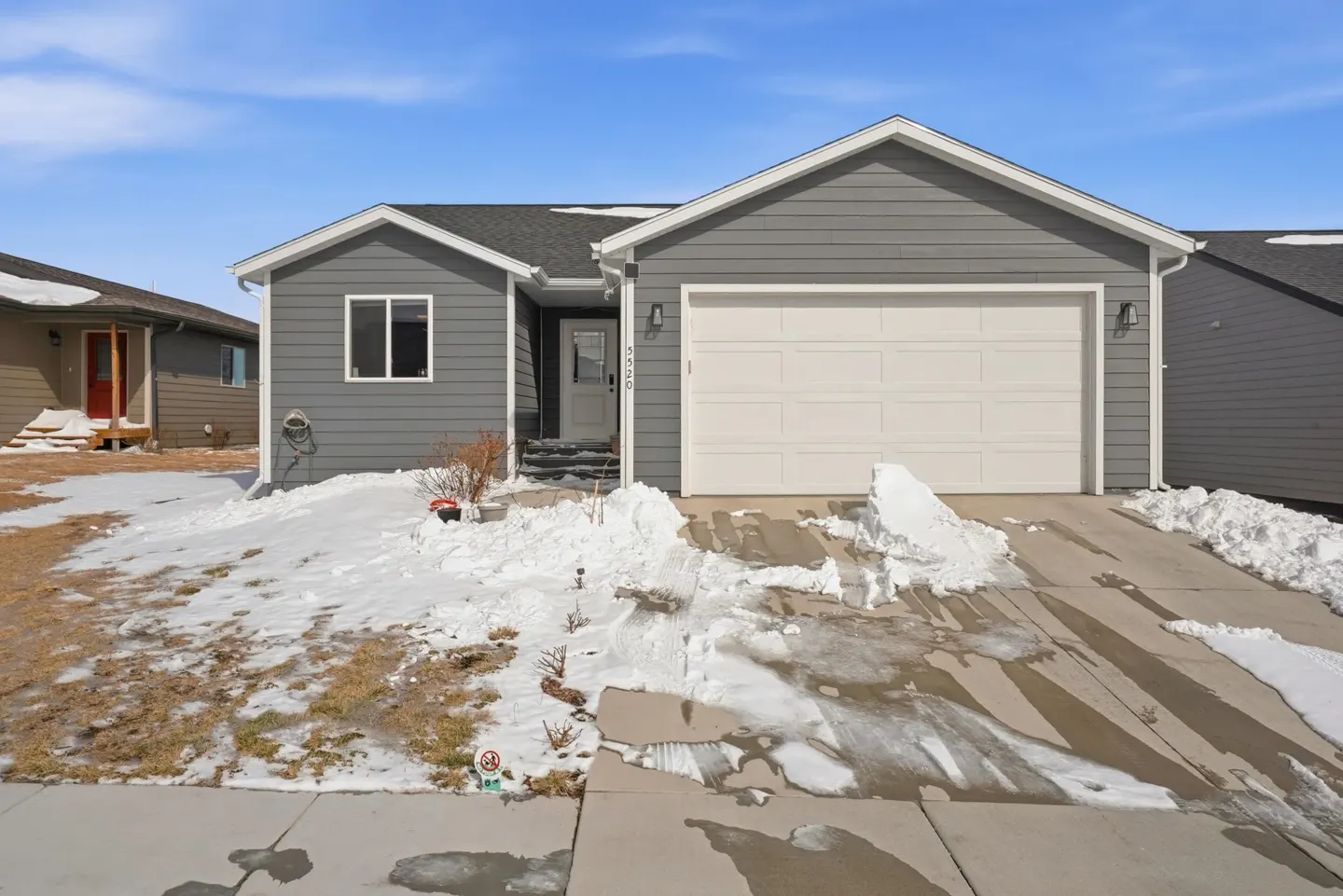 Single-story gray house with a white garage door and snowy front yard under a clear blue sky.