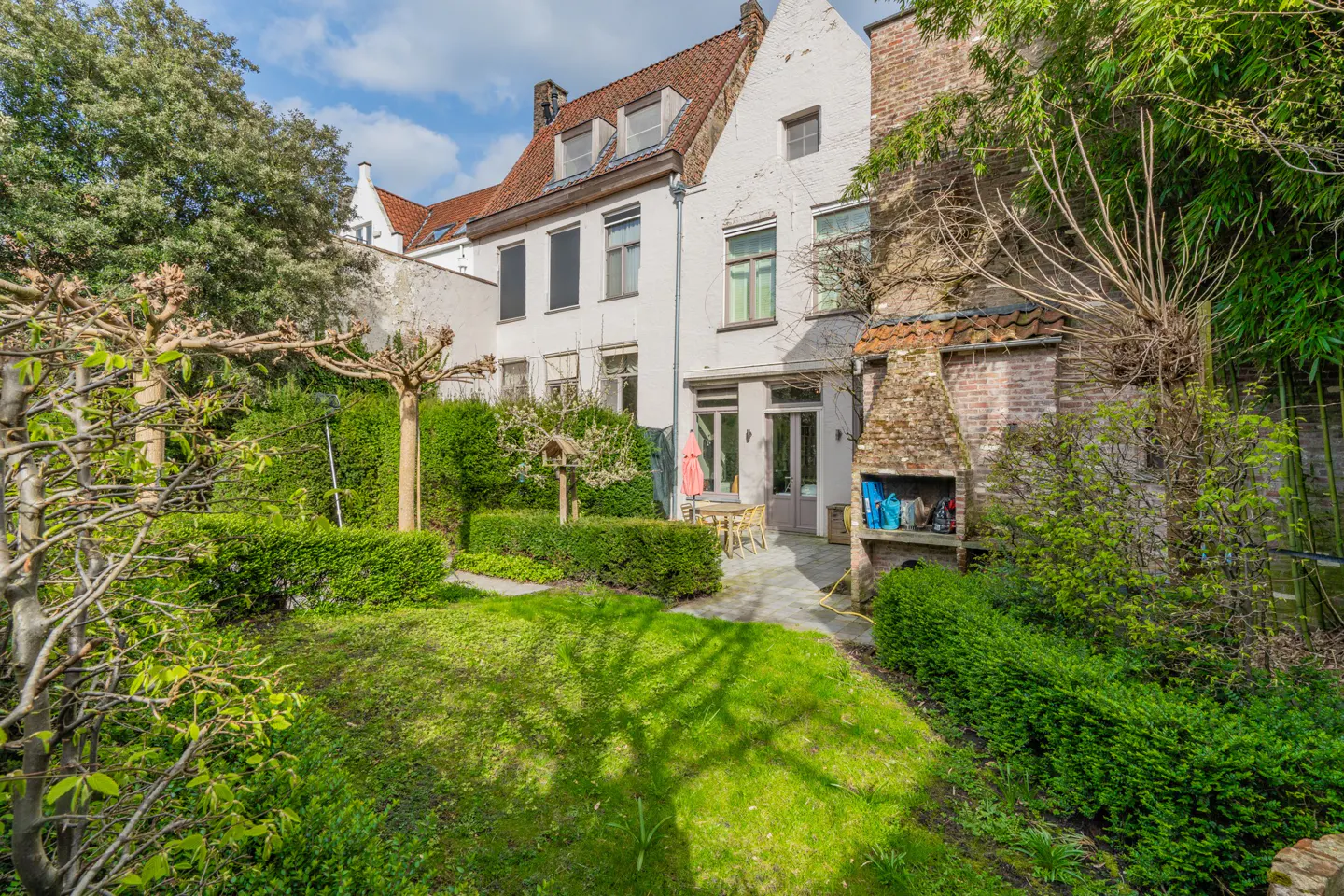 A lush green garden with a stone patio, outdoor fireplace, and a white multi-story house with a red tiled roof.