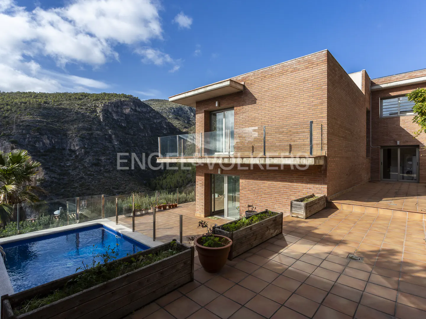 Exterior view of a modern brick house with a pool and mountain backdrop. The patio is tiled and has wooden planters. Blue sky with clouds.