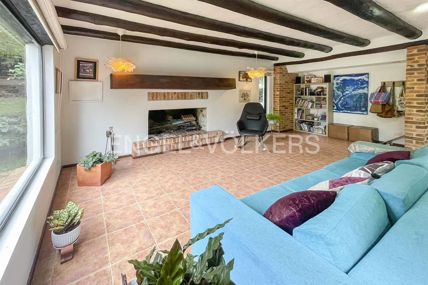 Living room with a blue sofa, brick fireplace, and dark wood beams on the ceiling. A large window lets in natural light.