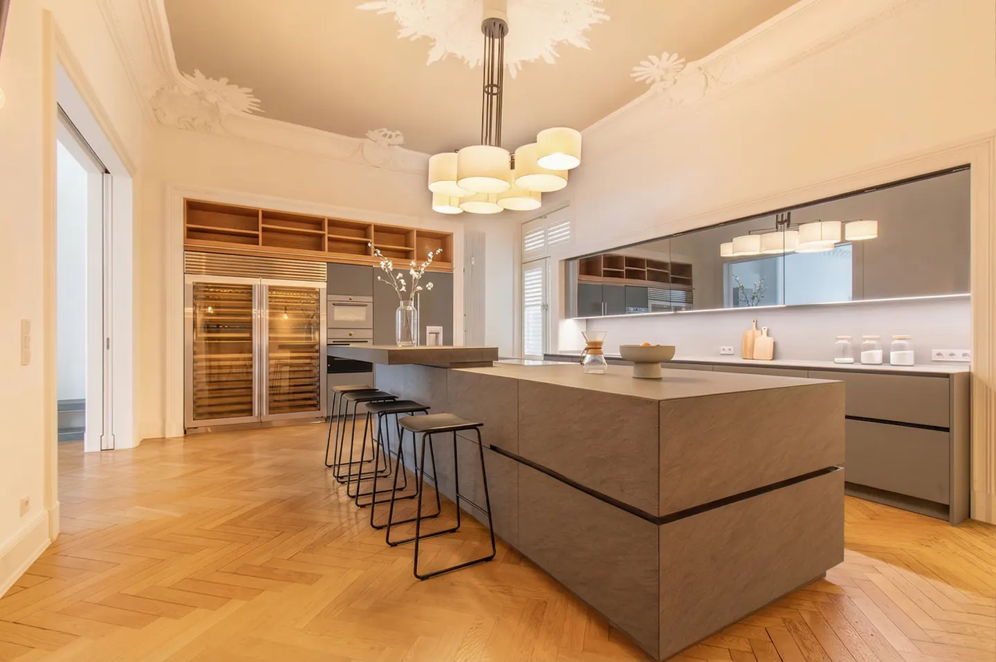 A modern kitchen with gray cabinets, a large island with black stools, and a herringbone wood floor. A wine fridge and decorative ceiling add luxury.