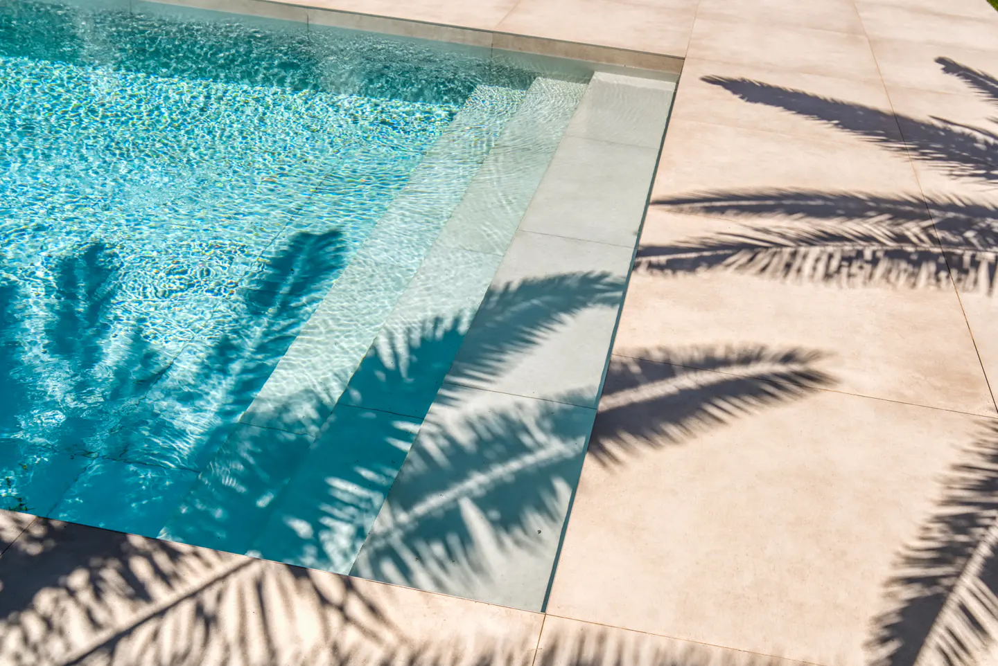 A sunlit swimming pool with steps and palm tree shadows on the beige tile surround.