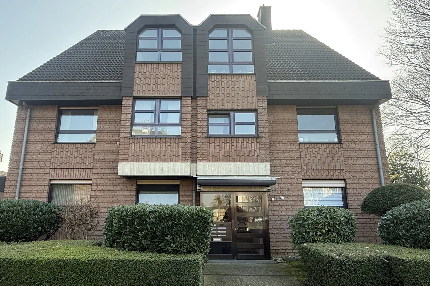 A two-story brick building with a dark roof and dark-framed windows. Green bushes line the front.