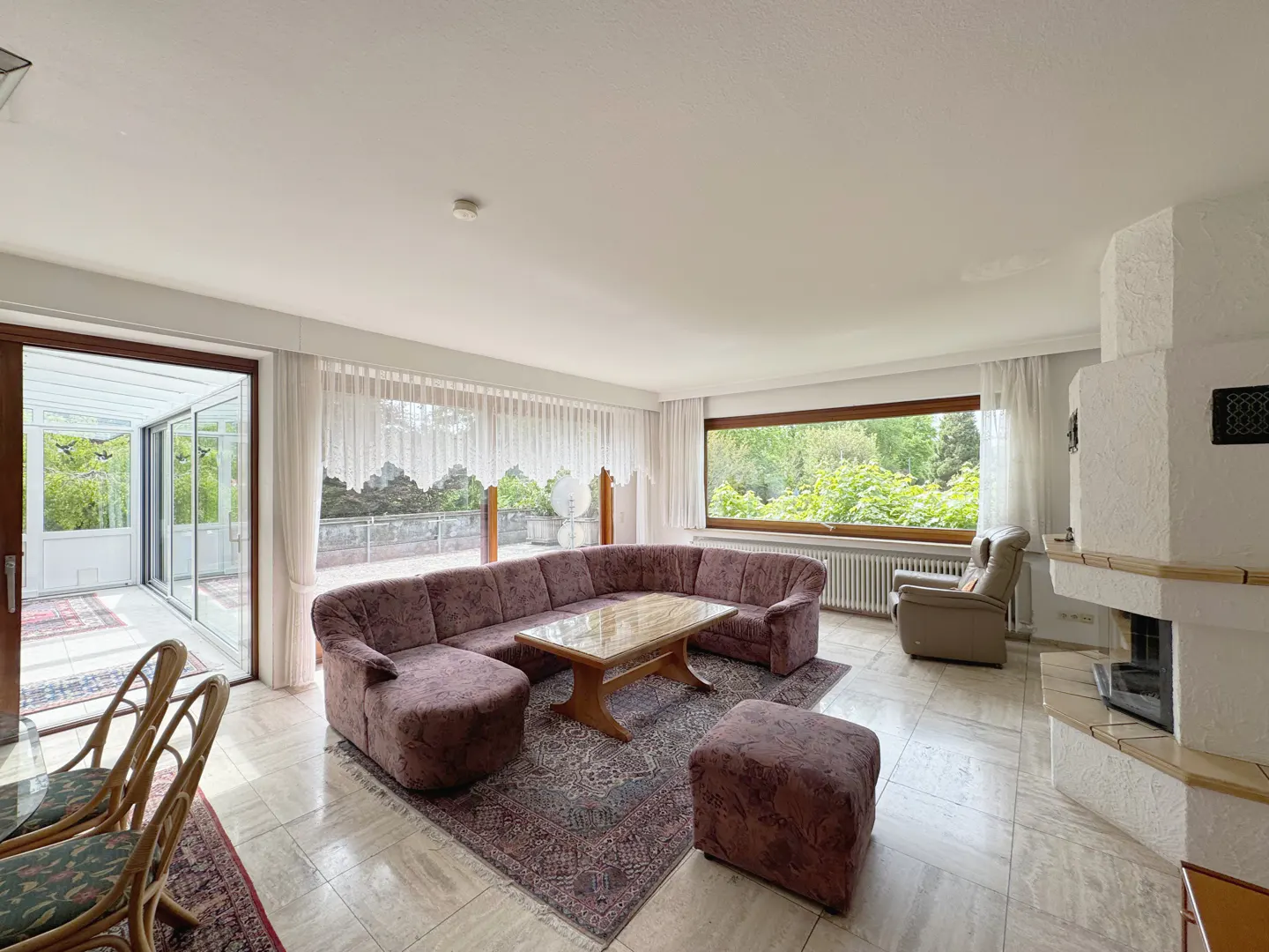 Bright living room with a large mauve sectional sofa, a wooden coffee table, and a white fireplace. A sunroom is visible through glass doors.