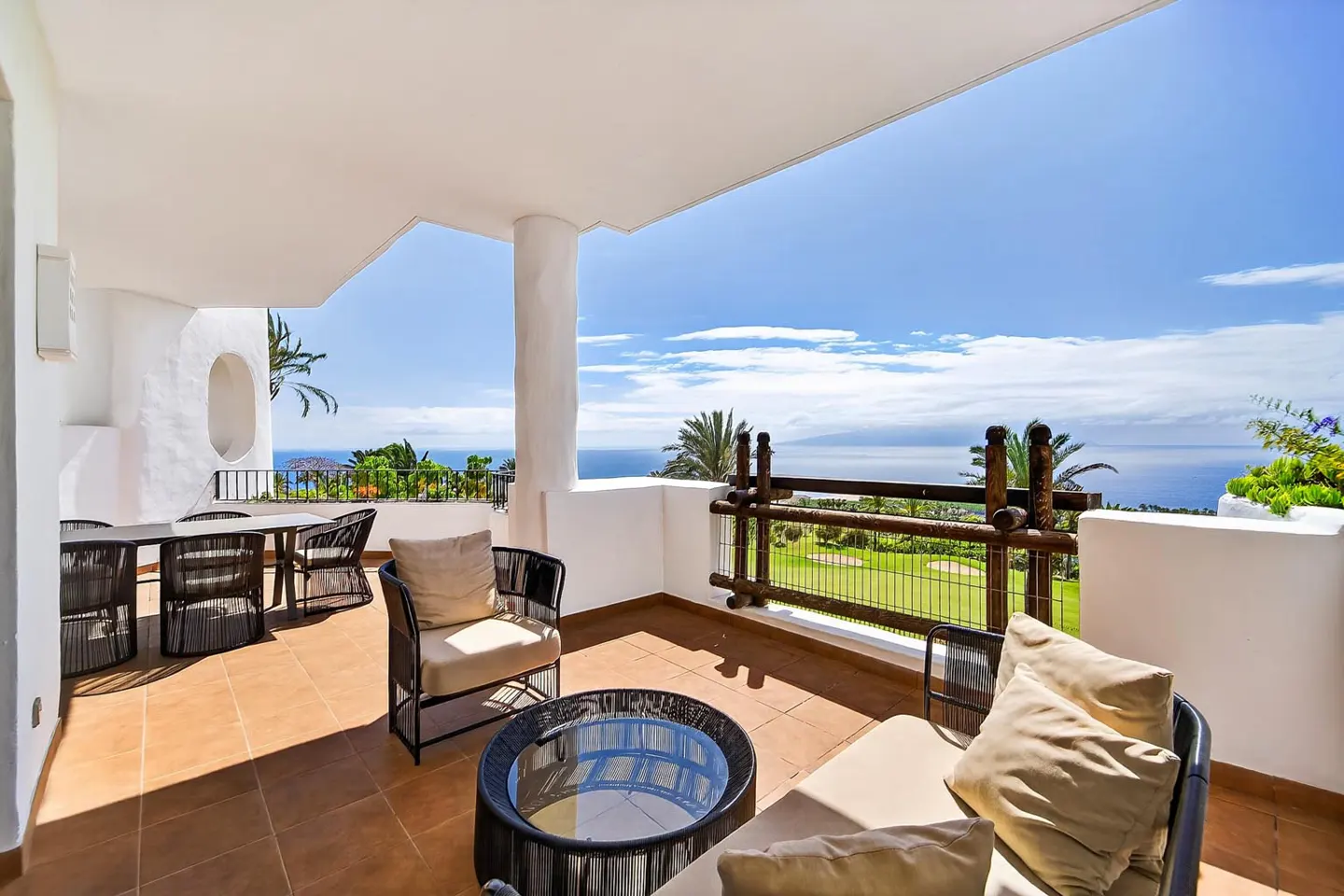 A sunny patio with seating overlooks a golf course and ocean view. White walls and a tiled floor create a bright, inviting space.
