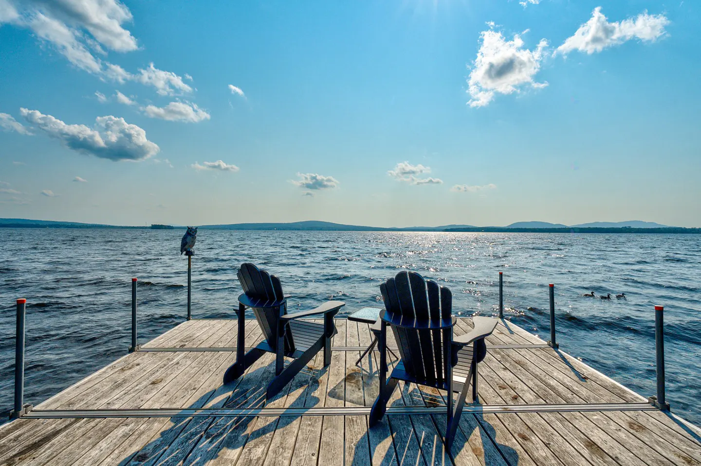 Two black Adirondack chairs face a lake from a wooden dock under a blue sky with scattered clouds.